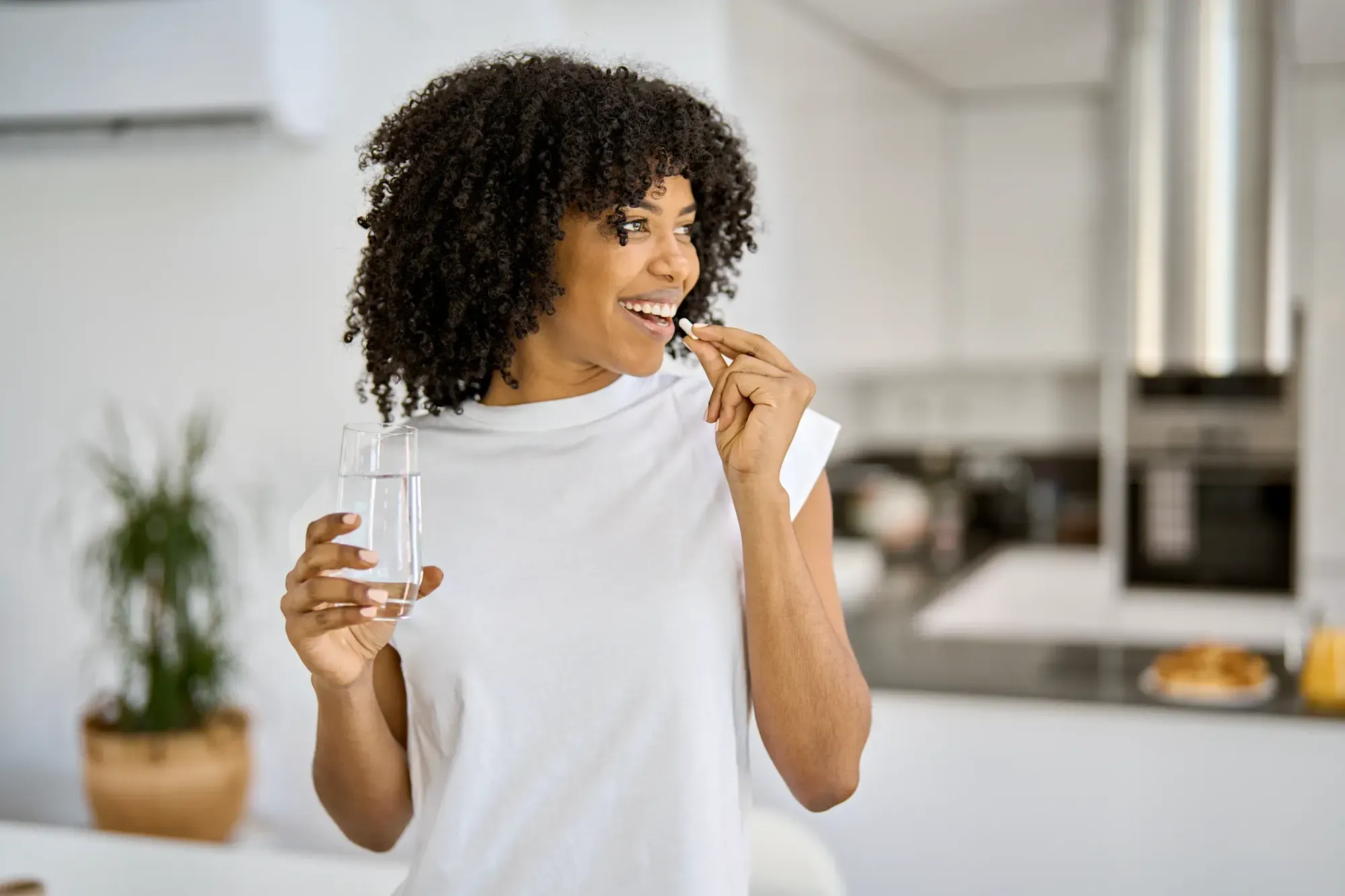 A smiling woman takes a FlowVeda capsule with a glass of water in a bright kitchen.